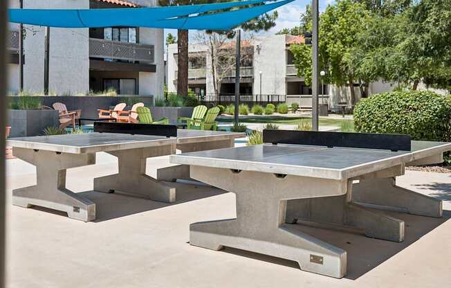 A concrete table with a blue canopy is in the foreground of a sunny outdoor area.