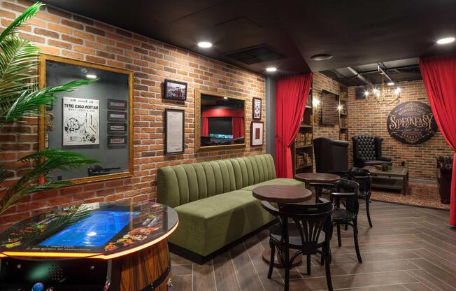 A stylish speakeasy-themed lounge with exposed brick walls, featuring a green velvet sofa, small round tables, and black leather armchairs. Red curtains frame the entrance, and a vintage pinball machine adds a playful touch to the cozy, elegant atmosphere. Subtle lighting enhances the inviting ambiance.