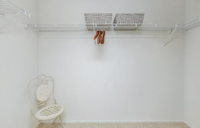 A spacious closet with white walls and a light-colored carpet. There are two wire shelves at the top holding two baskets, and a few wooden hangers are suspended from the shelf. A decorative metal chair with a woven seat is placed in one corner of the closet.