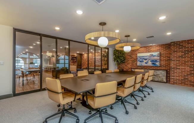 A modern conference room featuring a large rectangular table surrounded by ten ergonomic chairs. Soft lighting from pendant lamps illuminates the space, which has a brick accent wall. A large window allows natural light, and a separate meeting area is visible in the background with a plant and artwork.