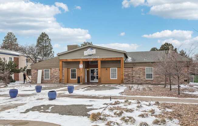 A building with a brown roof and a sign that says "LEASING CENTER" is surrounded by blue planters and snow on the ground.