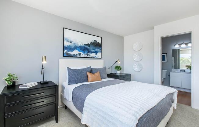 A modern bedroom featuring a gray and white color scheme. The bed is adorned with a textured white blanket and a gray throw, with decorative pillows. Two sleek black nightstands flank the bed, each with a small lamp and a potted plant. A framed abstract painting hangs above the bed, and an en-suite bathroom is visible in the background.