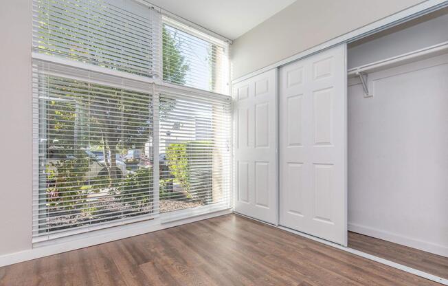 Bright room with large windows covered by white blinds, allowing sunlight to illuminate the space. The floor is wooden, and there is a sliding closet door on the right side. Outside, greenery is visible through the windows, adding a touch of nature to the interior.