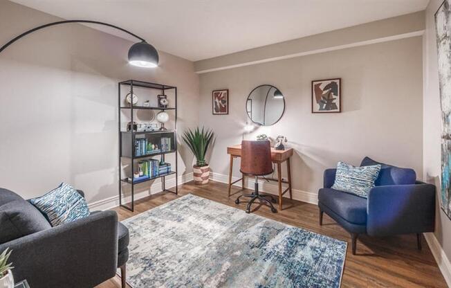 A living room with a grey couch, a blue couch, a rug, a desk, a chair, a potted plant, and a bookshelf.