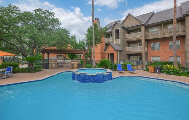 A vibrant pool area with a circular hot tub at the center, surrounded by lush greenery and palm trees. Comfortable lounge chairs are set around the pool, and a barbecue area is visible nearby. In the background, a multi-story apartment building is partially seen under a clear blue sky.