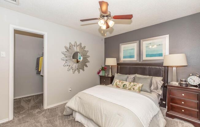 A cozy bedroom featuring a king-size bed with light-colored bedding and decorative pillows. There are two bedside tables with lamps and a round mirror on the wall. Above the bed, two framed artworks are displayed. A closet door is visible to the left, and the room has neutral-colored walls and carpet.