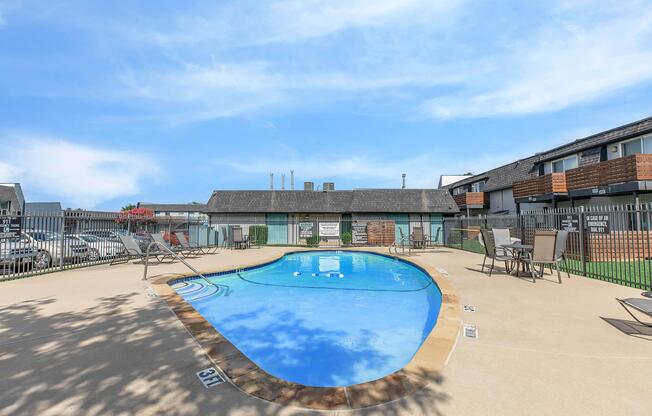 A clear blue swimming pool surrounded by a tan deck and lounge chairs, with a fenced area in a residential complex. In the background, there are buildings with a gray roof and some greenery, under a bright blue sky.