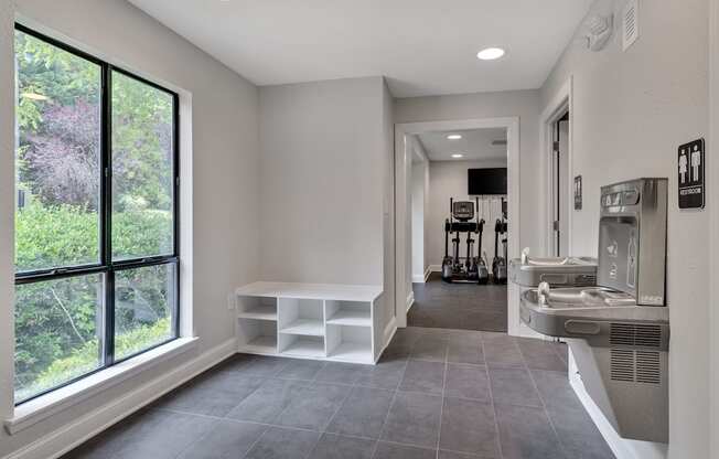 A room with a window, a bench, and water fountains outside of the fitness center.
