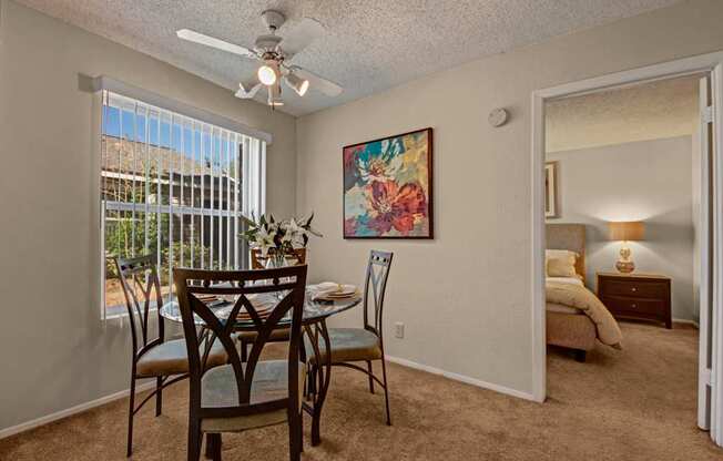 A bright dining area here at Cypress Pointe with beige carpet, neutral walls, and a large window with vertical blinds bringing in natural light. A ceiling fan adds comfort, while the open layout offers a clear view into the bedroom, creating a warm and inviting space for everyday dining.