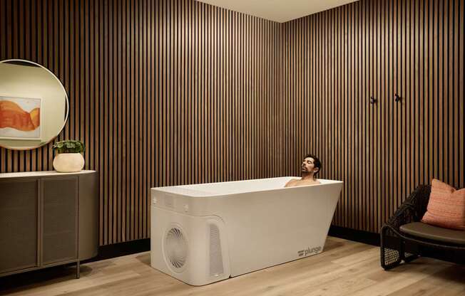 A man is sitting in a white bathtub in a room with wooden walls.