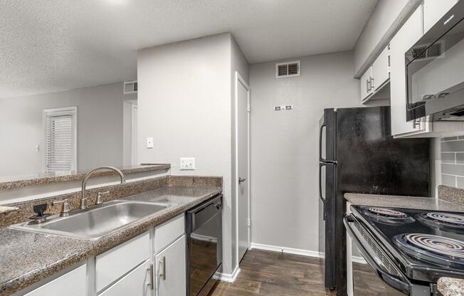 A modern kitchen featuring granite countertops, stainless steel sink, black appliances including a refrigerator and stove, and white cabinetry. The walls are painted a light gray, and there are wooden floors. The space is well-lit with overhead lighting, giving a clean and contemporary look.
