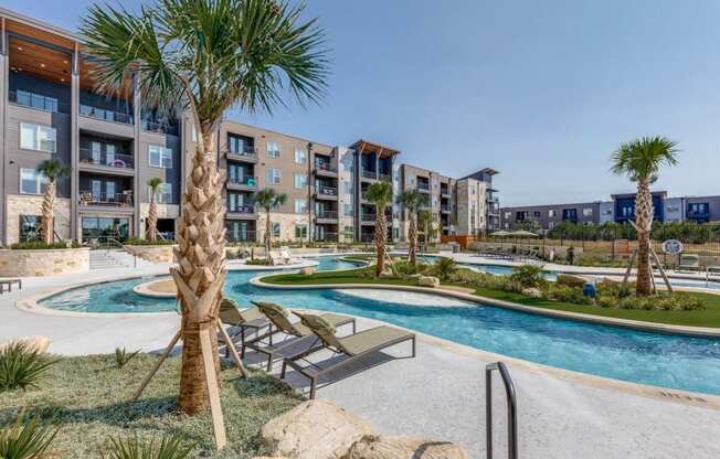 A pool surrounded by palm trees and lounge chairs in front of apartment buildings.