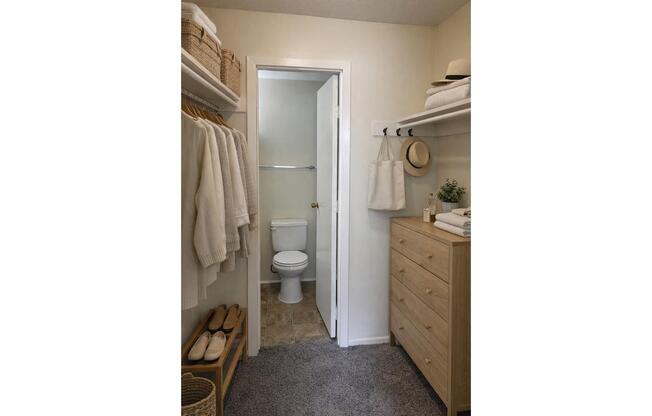 A neatly organized bathroom featuring a small closet space with hanging white towels, a toilet in the background, and a wooden storage unit with decorative items. Soft lighting highlights the clean, minimalistic decor, contributing to an inviting ambiance.