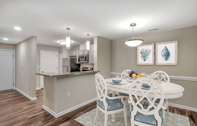 A virtually staged dining room with gray walls with white trim, hardwood style flooring and a pendant light in the center of the ceiling. A kitchen with granite countertops and stainless steel appliances is located in the background.