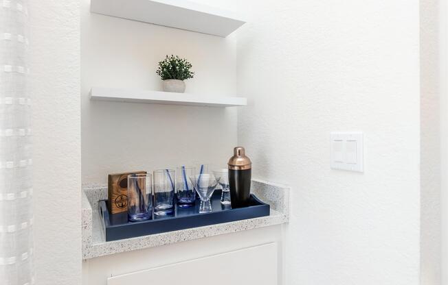 A home bar setup featuring a tray with various glassware, including tall and short glasses, a cocktail shaker, and a wooden box. Above, there are two white shelves with a small potted plant. The wall is a soft, light color, creating a clean and modern appearance.
