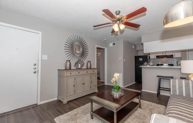 A cozy living room featuring a modern design. The space includes a light-colored sofa with striped patterns, a wooden coffee table, and a decorative area rug. A large wall mirror and silver vases add elegance. A ceiling fan with wooden blades and an open kitchen area are visible in the background.