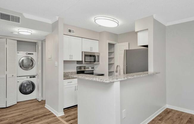 Modern kitchen area featuring white cabinets and stainless steel appliances, including a microwave and refrigerator. An island with a granite countertop separates the kitchen from the living space. A stackable washer and dryer are visible in the adjacent laundry area, with light wood flooring throughout.