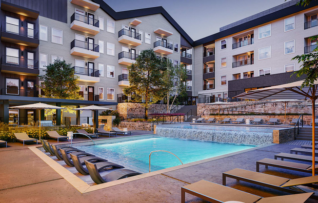 A swimming pool surrounded by lounge chairs and umbrellas in front of apartment buildings.