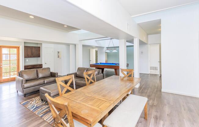 Interior view of a modern living space featuring a wooden dining table with chairs, two brown sofas, and a pool table in the background. The room has white walls, large windows allowing natural light, and a light wood floor. A doorway leads to the exterior area, enhancing the open feel of the space.