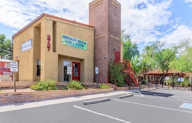 A two-story apartment building with a staircase on the side, featuring a beige exterior and a red door. A large sign on the front indicates "IMMEDIATE MOVE IN" with additional text. Surrounding the building are landscaped areas with shrubs and a parking lot visible in the foreground.