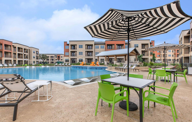 A table with green chairs is set up under a striped umbrella next to a pool.