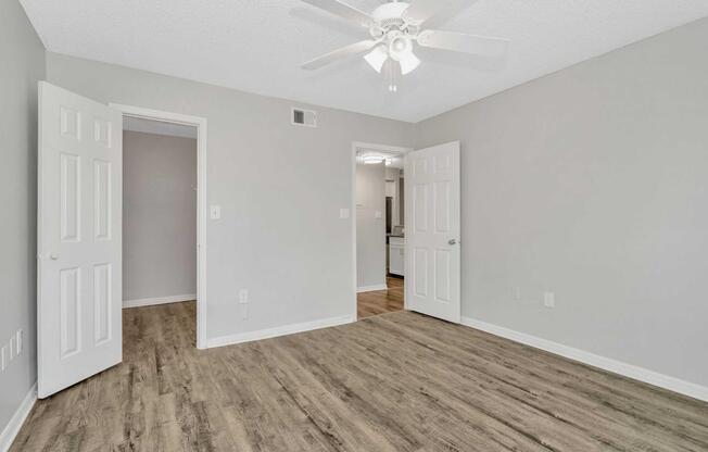 A spacious, empty room with light-colored walls and a ceiling fan. Two doors are open revealing small closets and other rooms. The floor features a wood-like laminate, creating a warm and inviting atmosphere. Natural light fills the space, enhancing its openness.