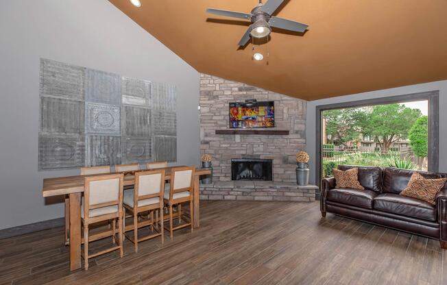 A spacious interior living area featuring a modern dining table with chairs, a stone fireplace, and a large window with outdoor views. The walls are painted in soft gray, and the ceiling is a warm orange. A flat-screen TV is mounted above the fireplace, and decorative elements include art pieces and throw pillows on the couch.