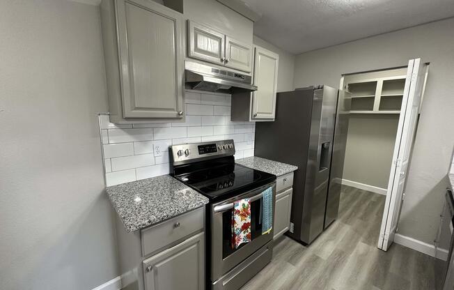A modern kitchen featuring gray cabinets, a stainless steel refrigerator, and a black electric stove with an oven. The countertops are granite, and there are white subway tiles as a backsplash. A colorful dish towel hangs from the stove. The flooring is light wood. A pantry space is visible to the right.