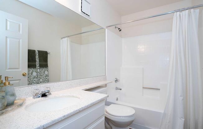 A clean bathroom featuring a sink with a countertop, a mirror, and a bathtub with a shower curtain. The walls are painted a light color, and there's a towel rack with neatly arranged towels. The overall appearance is modern and well-maintained.