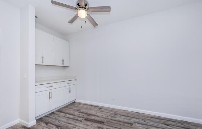 An empty room featuring light-colored walls, a ceiling fan with wooden blades, and a white cabinet with countertop. The floor is made of wood-like planks, creating a spacious and modern atmosphere. The room is well-lit, emphasizing its uncluttered design.