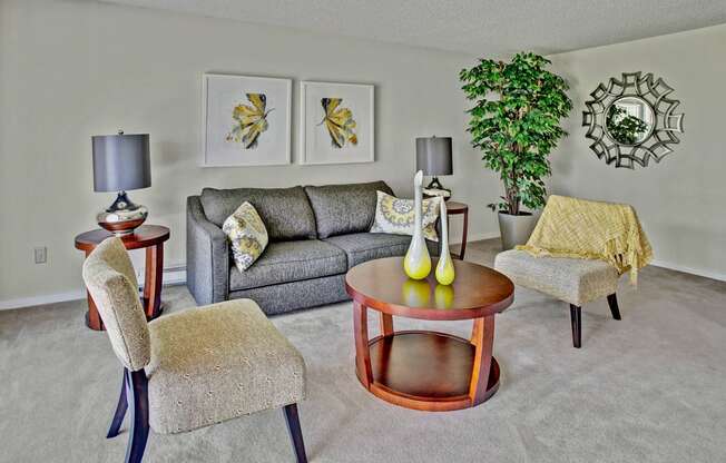 A welcoming living room here at Andorra featuring a plush gray sofa accented with patterned throw pillows, two upholstered accent chairs, and a round wood coffee table centered on soft carpeted flooring, complemented by matching table lamps, framed wall art, a decorative wall mirror, and a tall potted plant that adds warmth and visual interest.
