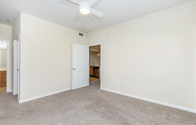 A spacious, neutral-colored room with a ceiling fan and light fixture, featuring beige carpeting. The room has one closed white door, leading to another area, and a doorway visible in the background, suggesting an open layout. Soft lighting enhances the inviting atmosphere.