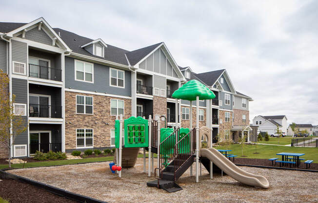 A playground with a green umbrella and slide in front of apartment buildings.