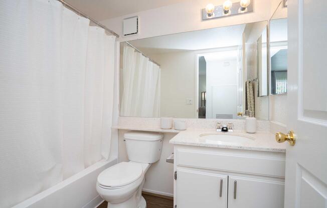 A clean, modern bathroom featuring a white shower curtain, a toilet, and a vanity with a mirror. The countertop is adorned with decorative items, and the lighting is provided by overhead fixtures. The walls are painted light colors, creating a bright and spacious feel.