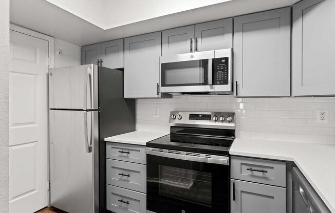 Modern kitchen featuring gray cabinetry, stainless steel appliances including a refrigerator, microwave, and oven. The countertops are white, complemented by a white tiled backsplash. The kitchen layout is efficient with ample storage and a door leading to another area.