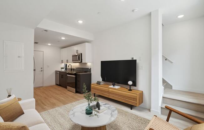 Modern open-plan living space with hardwood flooring, featuring a cozy sitting area with a white round marble-top coffee table, sleek TV stand, and an understairs white door.