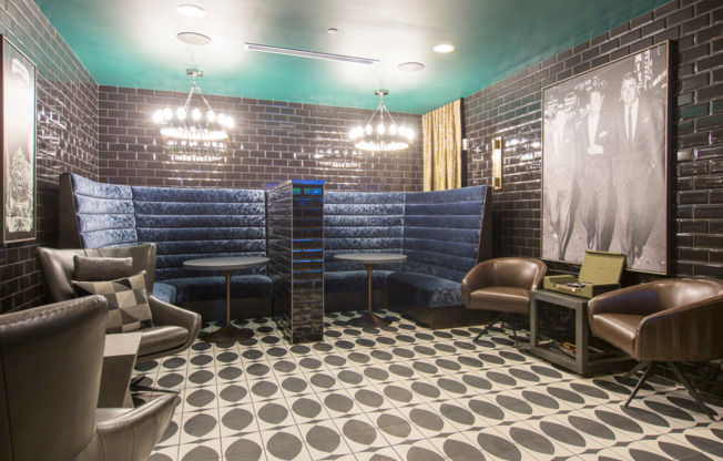 A seating area with two banquette-style booths, armchairs and side tables along the walls, dark subway-tile walls and moody lighting. There is a black-and-white vintage photo of three men in suits hung on the wall.