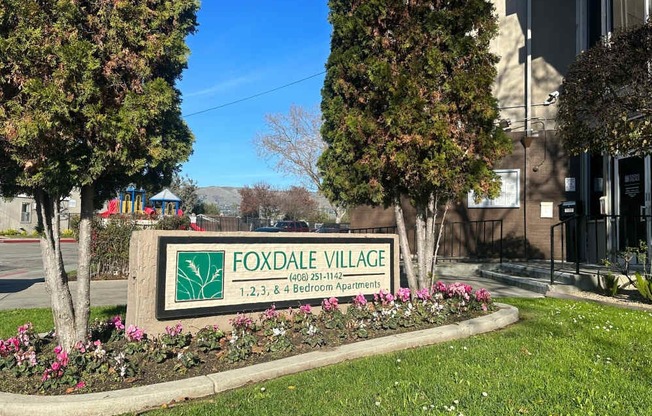 A sign for Foxdale Village with a phone number and address is surrounded by trees and flowers.