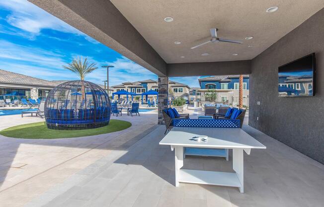 Outdoor lounge area featuring a shaded seating space with blue chairs, a white table, and a large round wicker-style chair. In the background, a swimming pool and several modern buildings are visible under a clear blue sky. The setting conveys a relaxed, resort-like atmosphere.