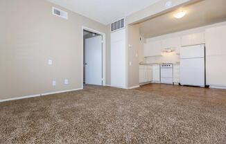 A view of an empty apartment interior featuring beige walls and light brown carpet. The space includes a white kitchen visible in the background with a refrigerator, stove, and cabinets. A doorway leads to another room, showcasing a clean and simple layout. Natural light illuminates the area.