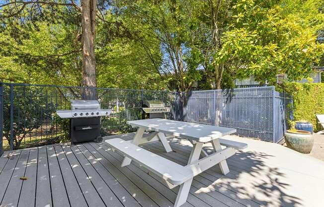 A picnic table is on a wooden deck with a grill and trees in the background.