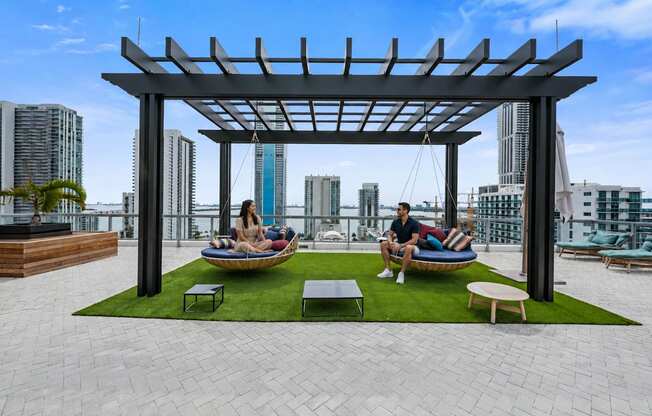 A man and woman are sitting on a couch under a pergola on a rooftop patio.