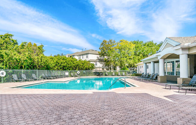 Swimming pool with large tanning deck, table with umbrella, lounge chairs, clubhouse in background surrounded by native landscaping