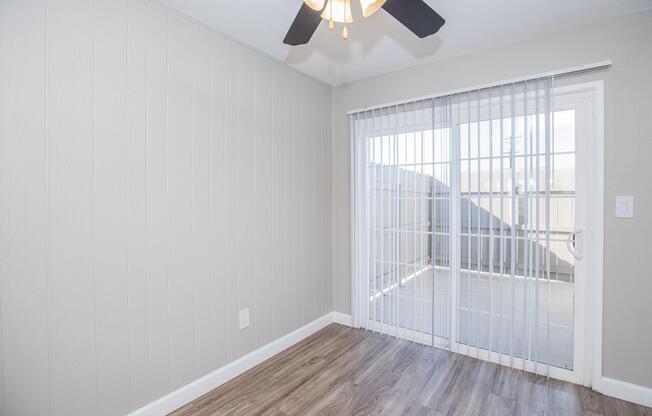 A bright, empty room featuring light gray walls and a ceiling fan. It has a sliding glass door leading to a balcony, with natural light streaming in. The floor is covered in light wooden laminate. The overall ambiance is clean and modern, suitable for various uses.