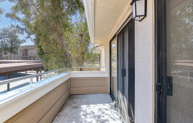 A balcony view with a smooth concrete floor, surrounded by a low railing. Lush greenery is visible in the background, providing a natural setting. A light fixture is mounted on the wall, and sliding glass doors offer access to the interior space. The atmosphere is bright and inviting.