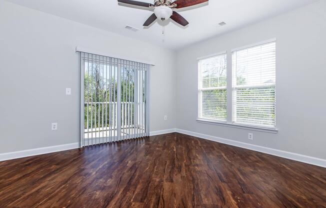 A spacious room with brown hardwood flooring, light gray walls, and a ceiling fan. Large windows provide natural light, and there is a sliding glass door leading to a balcony or patio area. Vertical blinds are partially drawn on the door, creating an open and airy atmosphere.
