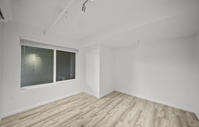 A blank, unfurnished room with light-colored walls and a wooden floor. There is a large window on one wall allowing natural light to enter, and a ceiling with exposed ductwork. The space appears modern and minimalistic, suitable for various uses or future decoration.