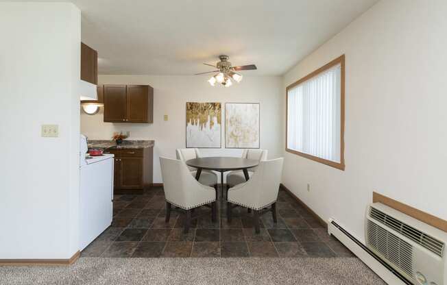 Fargo, ND Islander Apartments. A dining area with a four chair table that has a cailing fan with a light above it. A window brightens the room with natural light. Theres also a kitchen in the background