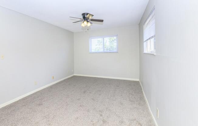 A bright, empty room with light gray walls and plush beige carpet. A ceiling fan with four light bulbs hangs from the ceiling. There is a window on one wall allowing natural light to enter. The space is clean and uncluttered, creating an inviting atmosphere.