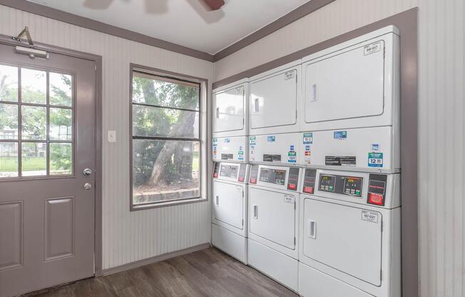 A laundry room featuring several stacked white washing machines and dryers against a wall, with a door leading outside and a large window letting in natural light. The floor is wooden, and there are trees visible through the window.